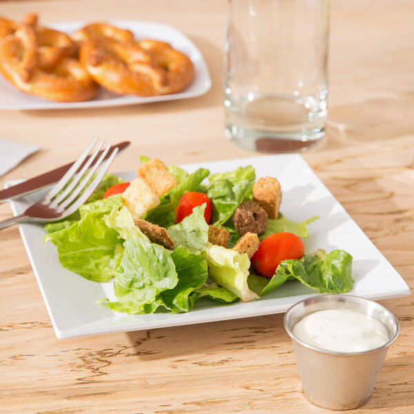 A white Siciliano square plate with salad and a fork on a table.