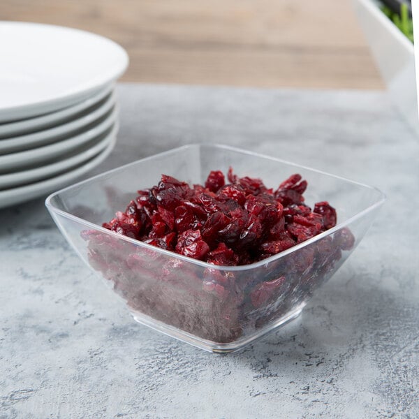 A clear small square acrylic bowl of cranberries on a table next to a stack of white plates.