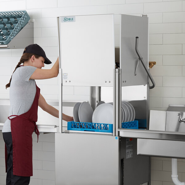 A woman in a red apron using the Noble Warewashing high temperature dishwasher to wash dishes.