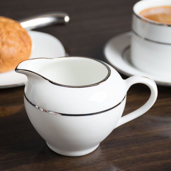 A 10 Strawberry Street white porcelain creamer with double silver lines on the bottom, sitting on a white saucer with a cup of coffee.