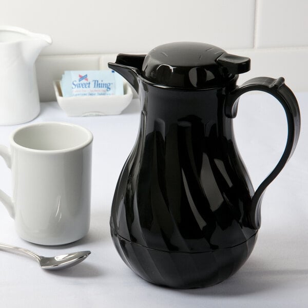 A black Choice coffee carafe on a table with a white mug.