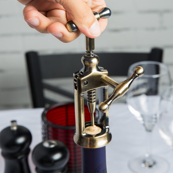 A person using a Franmara rack and pinion corkscrew to open a bottle on a white table with a black border.
