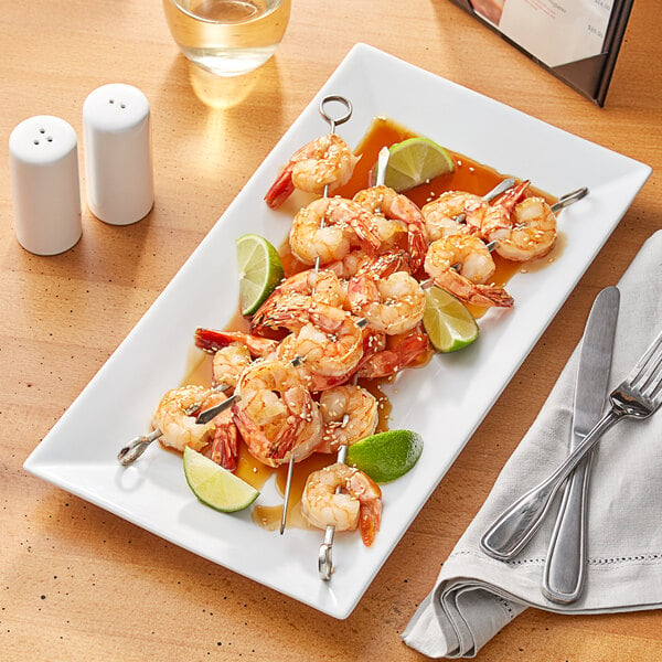 An Acopa bright white rectangular porcelain platter with shrimp skewers and lime wedges on a table.