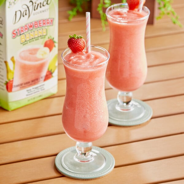 Two glasses of pink strawberry banana smoothies with straws and a carton of DaVinci Gourmet Strawberry Banana Real Fruit Smoothie Mix.