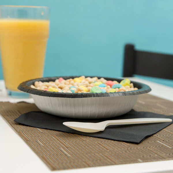 A bowl of Creative Converting black paper bowl filled with cereal with colorful candies on a table with a glass of juice.