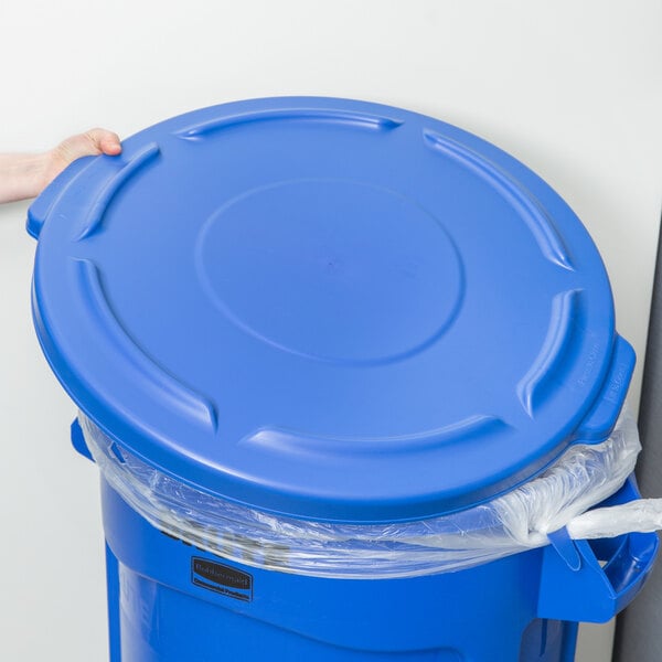 A person holding a blue Rubbermaid Brute trash can lid over a blue Rubbermaid Brute trash can.