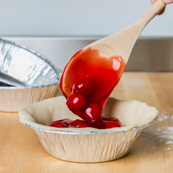 A spoon pouring red cherry pie filling into a D&W Fine Pack round foil pie tin.