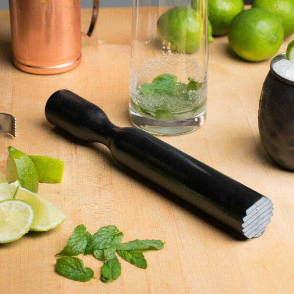 A close up of a black Franmara muddler with a ridged head being used to muddle limes and mint leaves in a glass.