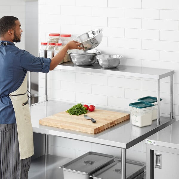 A man in a blue apron standing in a professional kitchen with a Regency stainless steel table mounted overshelf.