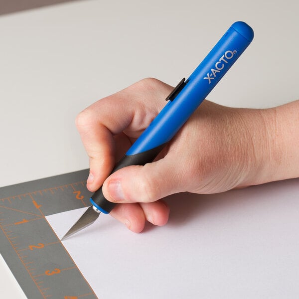 A hand holding a blue and black X-Acto Retract-A-Blade knife and cutting a piece of paper.