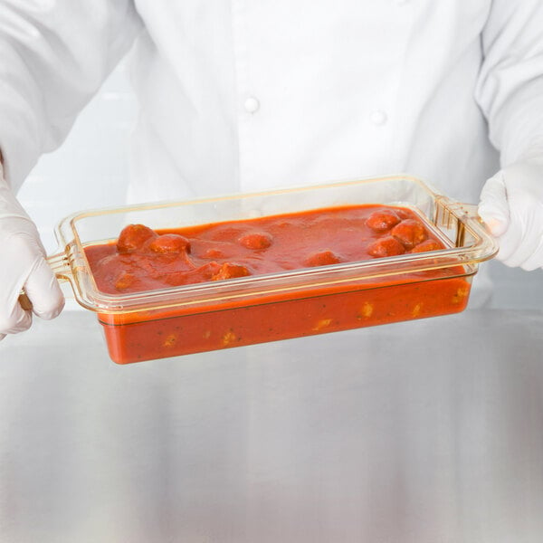 A person in white gloves holding a Cambro amber plastic food pan with red sauce on a counter.