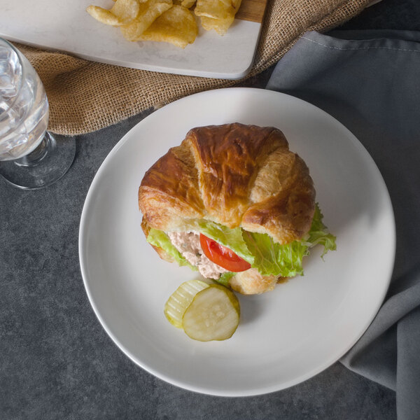 A croissant sandwich with lettuce, tomato, and pickles on a white GET Settlement melamine plate with a glass of water.