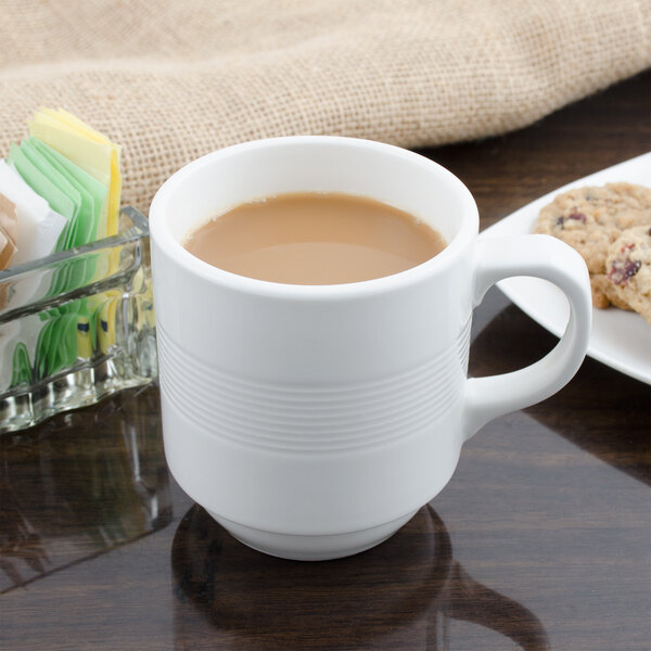 A white Bon Chef mug filled with brown liquid sitting on a table with cookies.