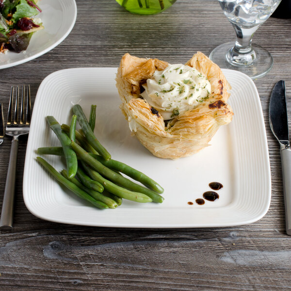 A white porcelain square plate with a pastry and green beans on it.