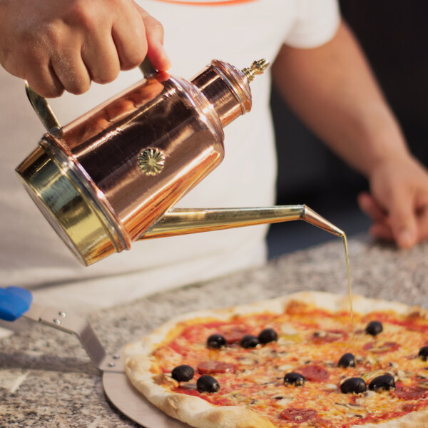 A hand pouring olive oil from a copper and brass cruet onto a pizza.