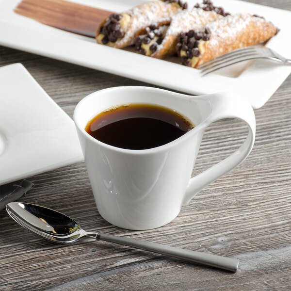 A white bone china cup of coffee on a table with a plate of pastries and a silver spoon.