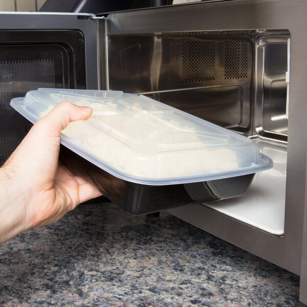 A hand placing a Pactiv Newspring VERSAtainer rectangular plastic container of food in a microwave.
