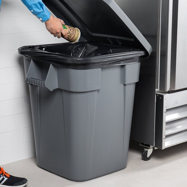 A man putting a coffee cup into a Rubbermaid BRUTE 40 gallon square trash can in a school kitchen.