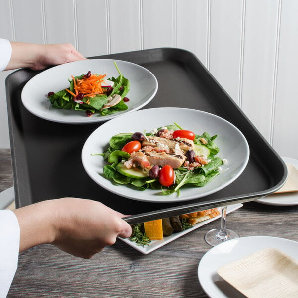 A person holding a Cambro black non-skid serving tray with a plate of salad on a table.