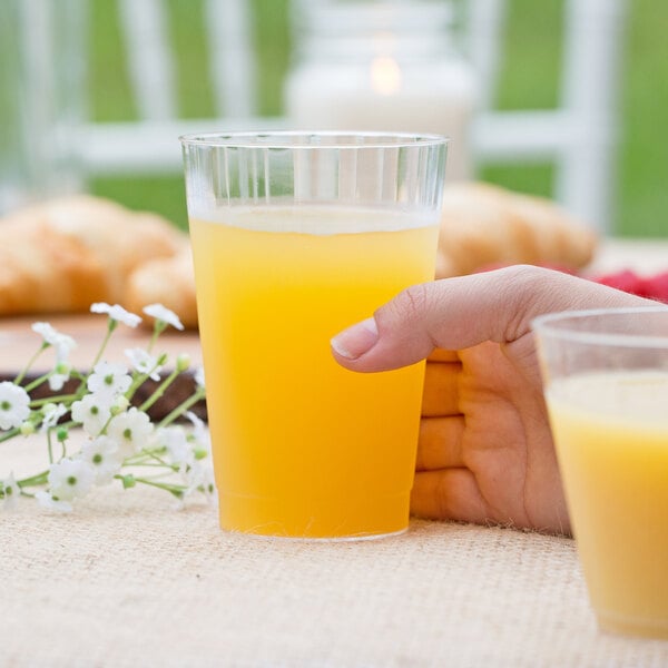 A person holding a WNA Comet Classicware tall clear plastic fluted tumbler filled with orange juice.