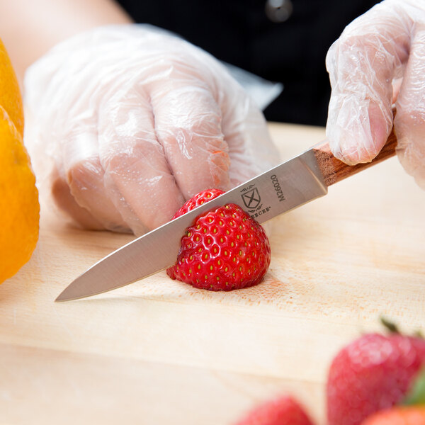 A person's hand in a plastic glove using a Mercer Culinary Praxis paring knife to cut a strawberry.