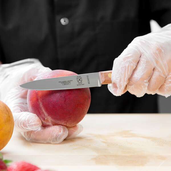 A person in gloves uses a Mercer Culinary Praxis paring knife to cut a peach.