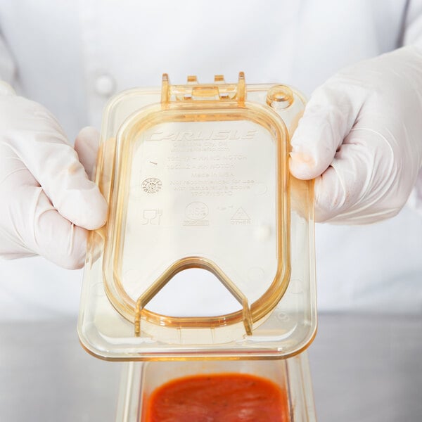 A gloved hand using a Carlisle Amber High Heat Hinged Lid to cover a plastic container with sauce.