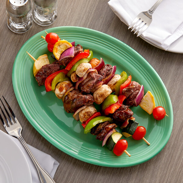 A Tuxton cilantro green oval china platter with skewers of meat and vegetables on a table.