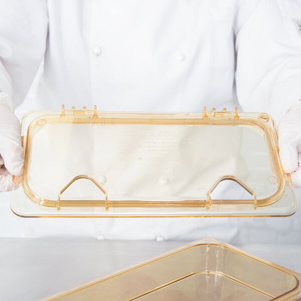 A person in white gloves holding a clear plastic container with a Carlisle Amber High Heat Hinged Lid.