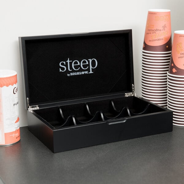 A black Steep by Bigelow tea chest with the lid open and a stack of white and brown paper cups inside.