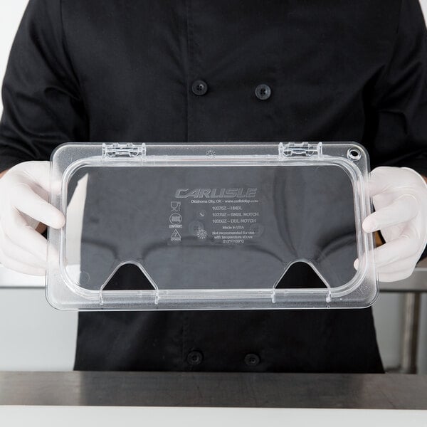 A person in a chef's uniform holding a Carlisle clear plastic hinged lid over a clear plastic container.