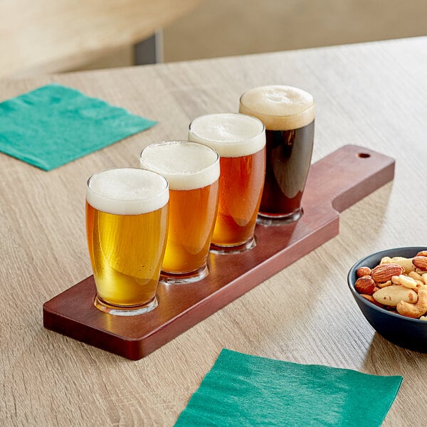 A mahogany flight paddle with Barbary tasting glasses filled with beer and a bowl of mixed nuts on a table.