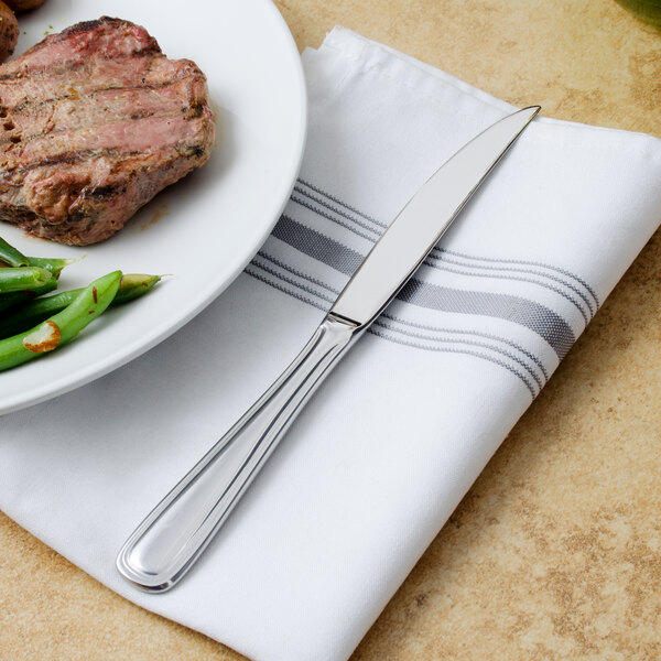 A Walco stainless steel steak knife on a napkin next to a plate of meat and green beans.
