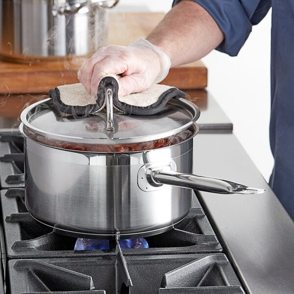A person wearing a glove holds a Vigor stainless steel sauce pan over a pot of food.