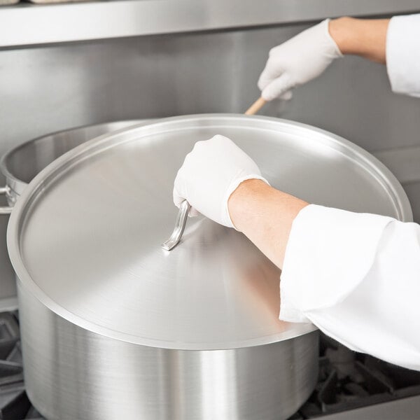 A person wearing gloves holding a Vigor stainless steel lid over a large silver pot.