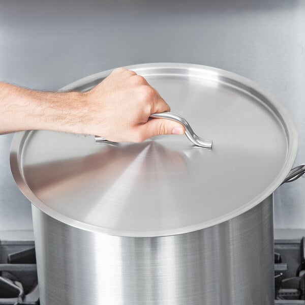 A hand holding a Vigor stainless steel lid over a large pot.