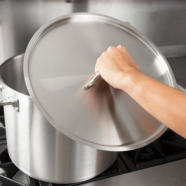A hand holding a Vigor SS1 Series stainless steel lid over a large pot on a stove.