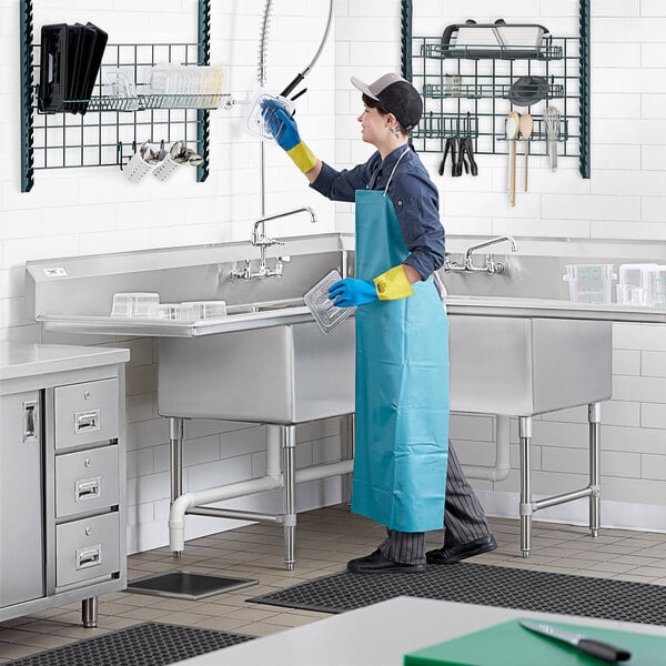 A woman in a blue apron and gloves cleaning a Regency stainless steel three compartment sink with drainboards in a professional kitchen.