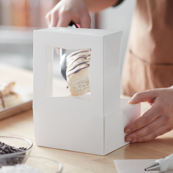 A person holding a piece of pie in a white Baker's Mark bakery box with a clear window.