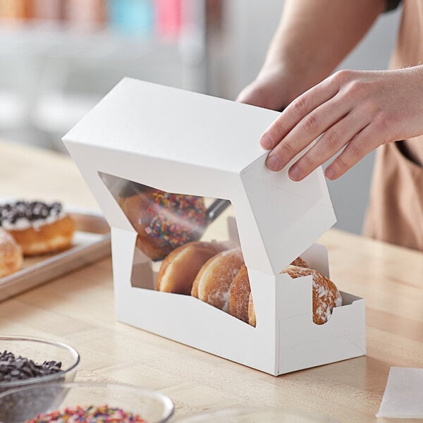 A person's hand holding a white Baker's Mark bakery box with a window showing donuts inside.