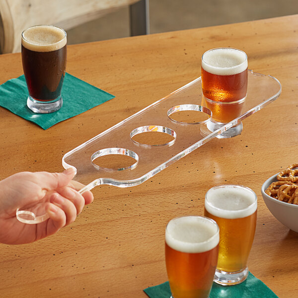 A hand holding an Acopa transparent plastic flight paddle with beer glasses on a table.