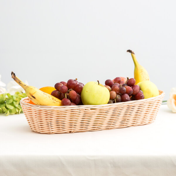 A Tablecraft woven rattan-like bread basket filled with bananas, apples, and fruit on a table.