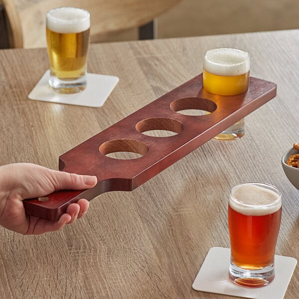 A hand holding an Acopa mahogany finish flight paddle with beer glasses on a table.