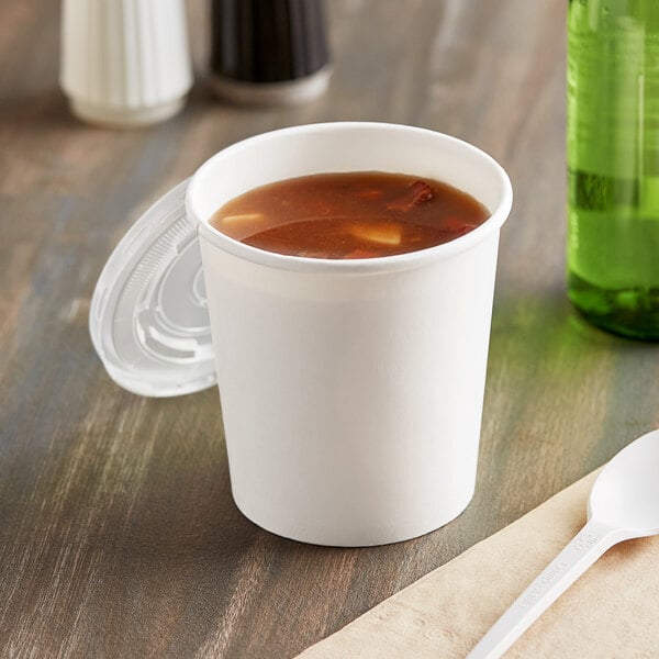 A white paper cup filled with soup and covered with a clear plastic lid on a table.