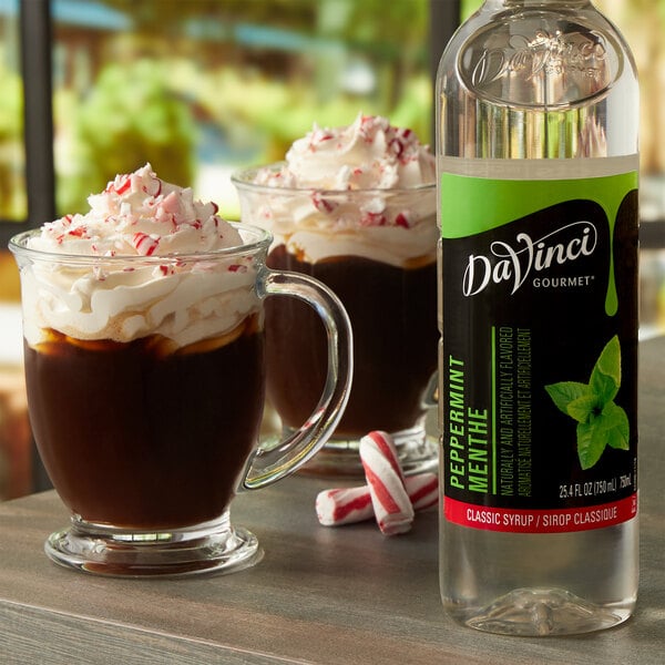 A bottle of DaVinci Gourmet Classic Peppermint Flavoring Syrup on a counter next to a glass mug of hot chocolate with whipped cream.