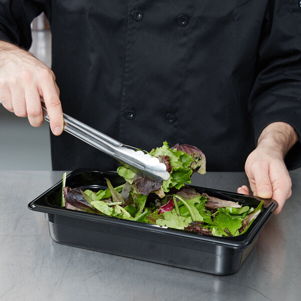 A person using metal tongs to serve salad from a Cambro black polycarbonate food pan.