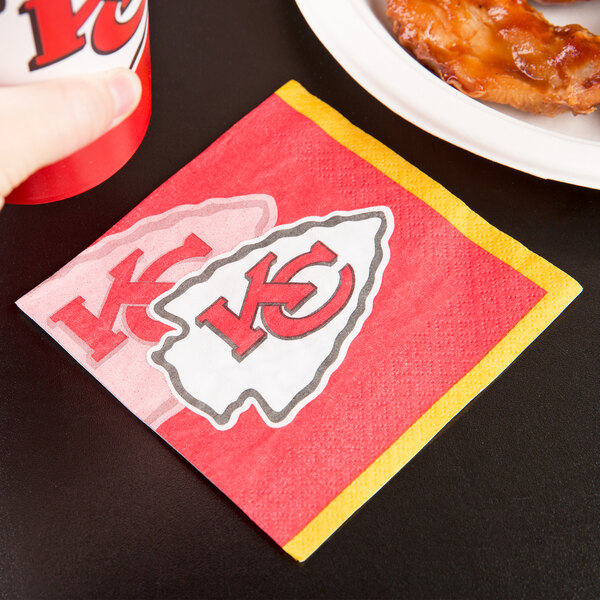 A Kansas City Chiefs beverage napkin with the team logo next to a plate of chicken.