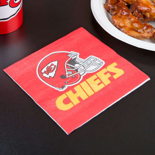 A red and white Kansas City Chiefs luncheon napkin with a helmet on it on a table next to a plate of chicken wings.