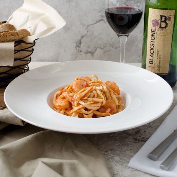 A white porcelain deep plate with pasta and shrimp on a table with a glass and bottle of wine.