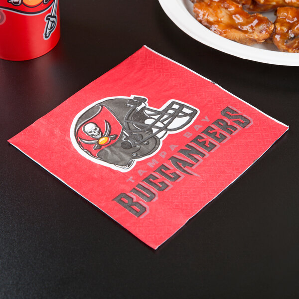 A red Tampa Bay Buccaneers luncheon napkin with a logo on it next to a plate of chicken wings.